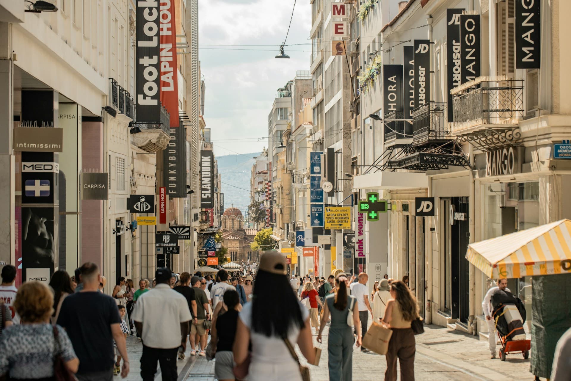 A crowded pedestrian shopping street in central Athens lined with retail signage