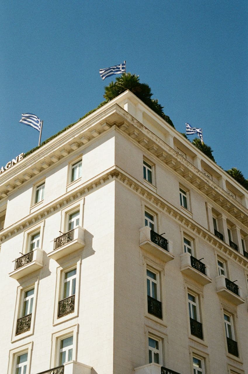 The corner facade of a historic Athens hotel with Greek flags on its rooftop terrace