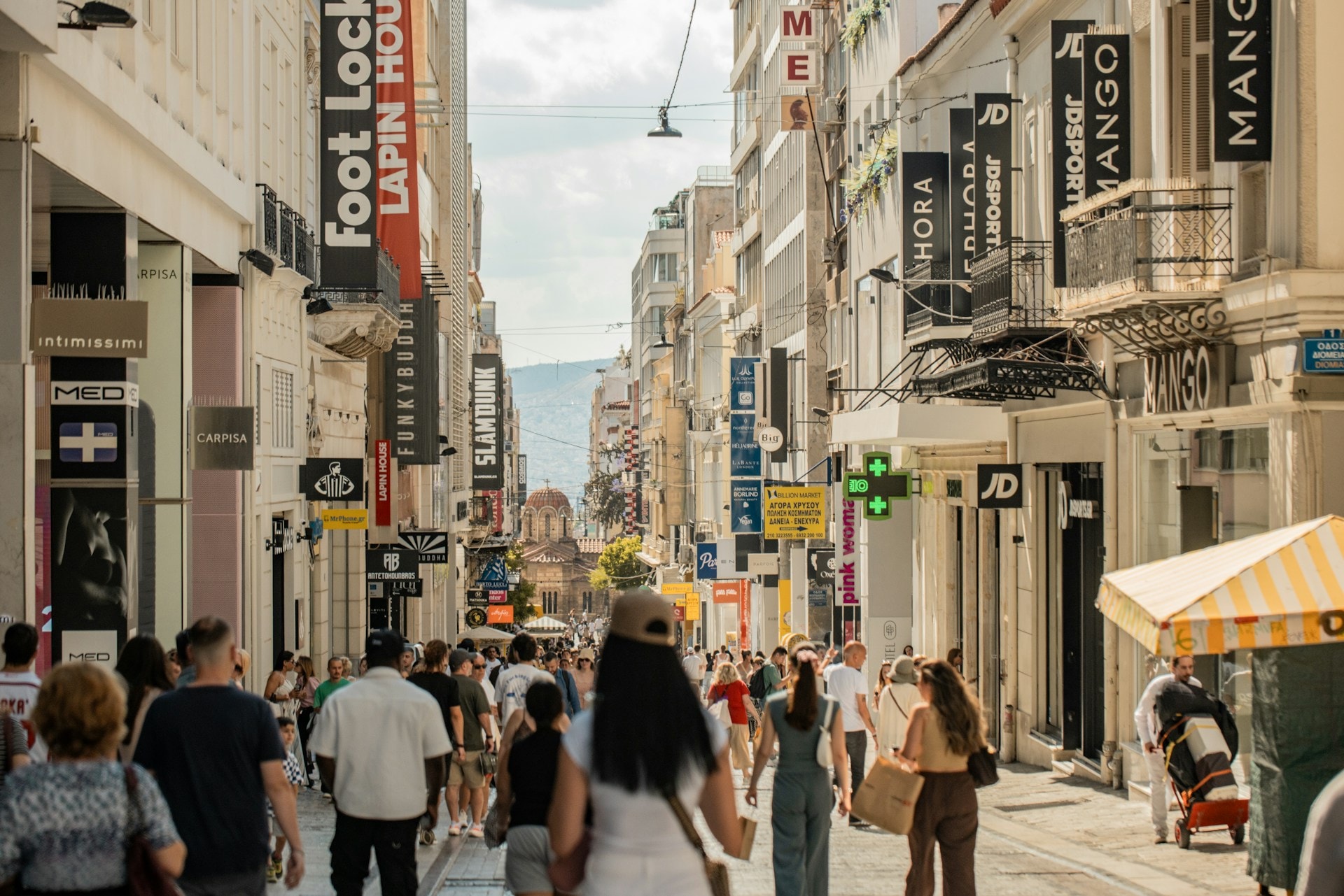 A crowded pedestrian shopping street in central Athens lined with retail signage